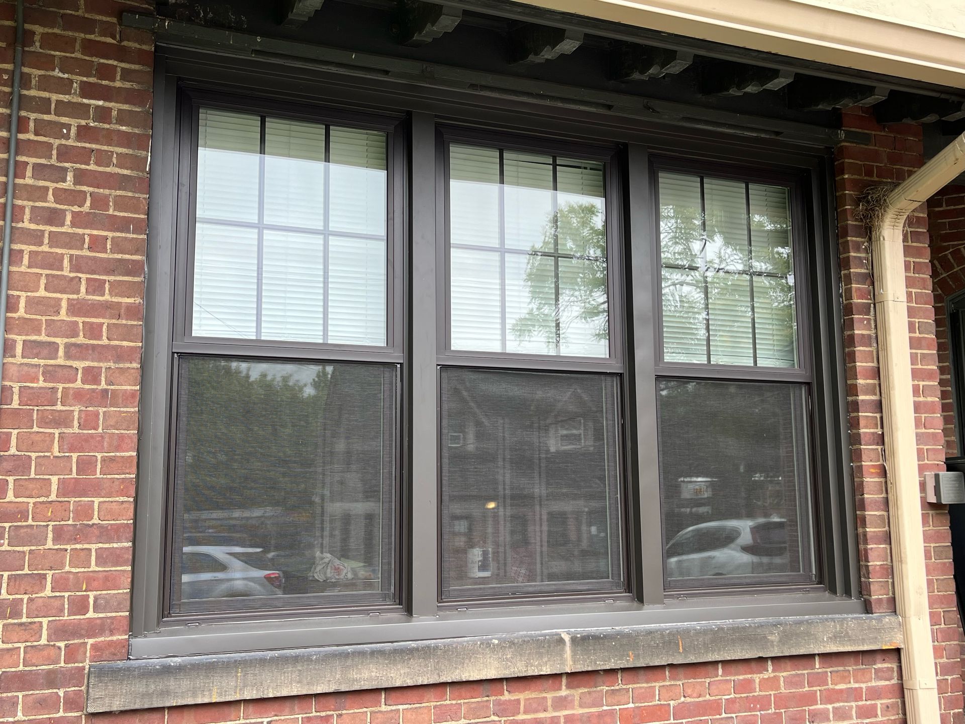 Brown-framed triple window with screens, set in a red brick wall. Blinds are visible through the glass.