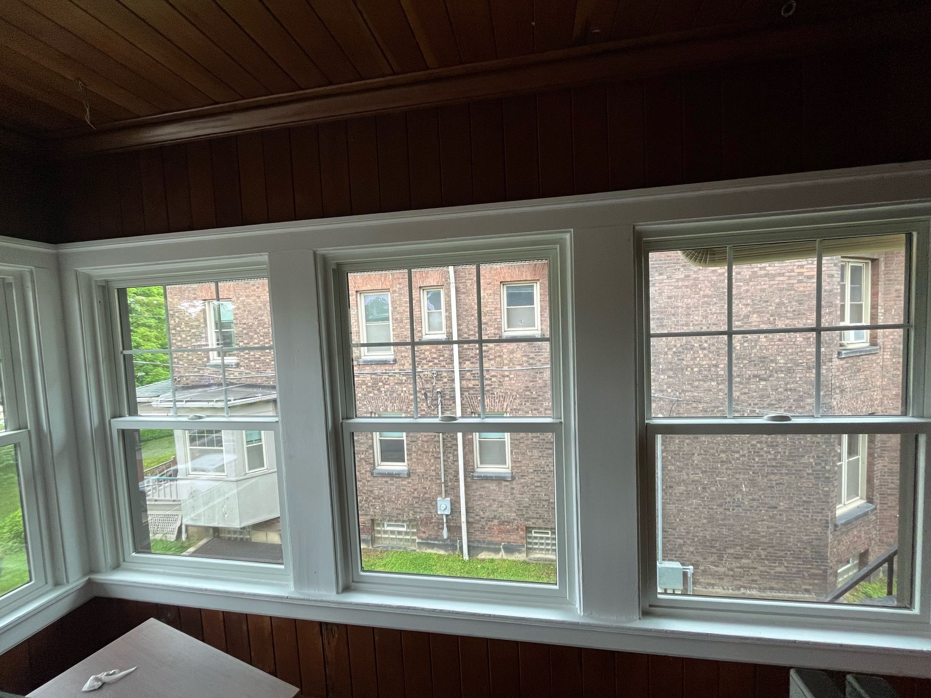 Three white-framed windows with views of a brick building. The interior has brown paneling and a dark ceiling.