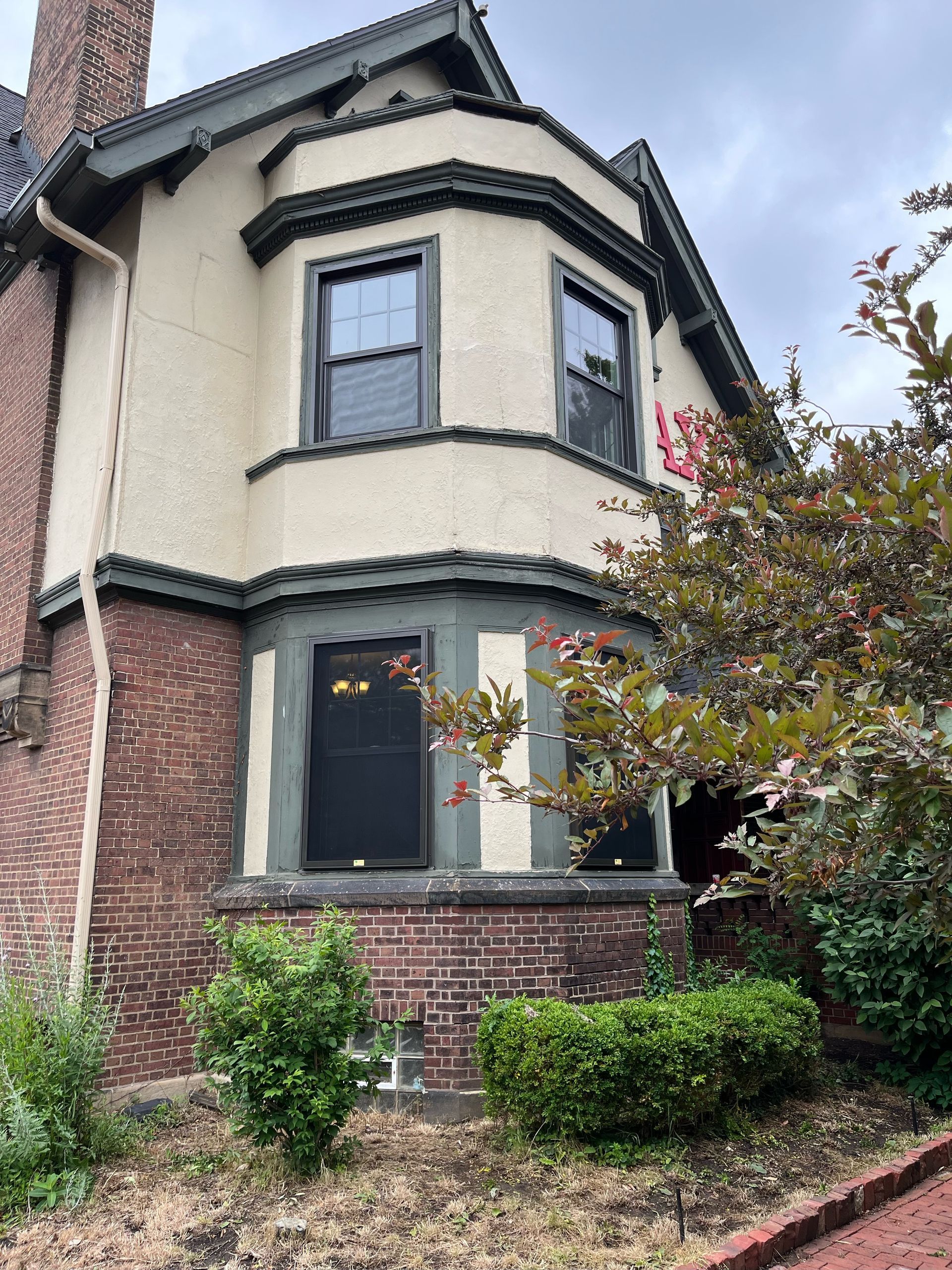 Two-story house with brick base, stucco upper, bay window, and dark trim. Green and brown foliage.