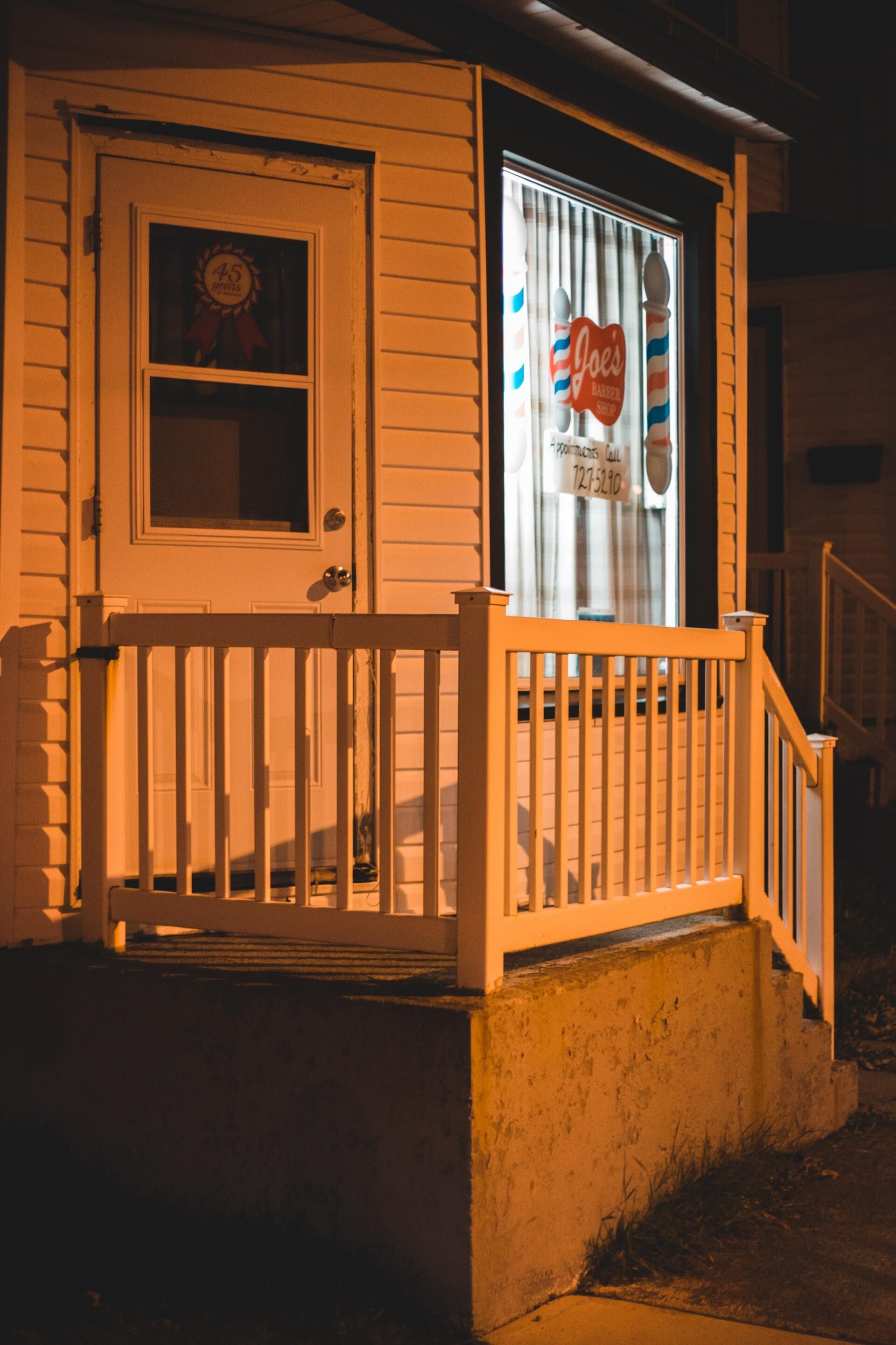 Exterior of a barbershop at night, illuminated, with a white door, and barber pole visible in window.