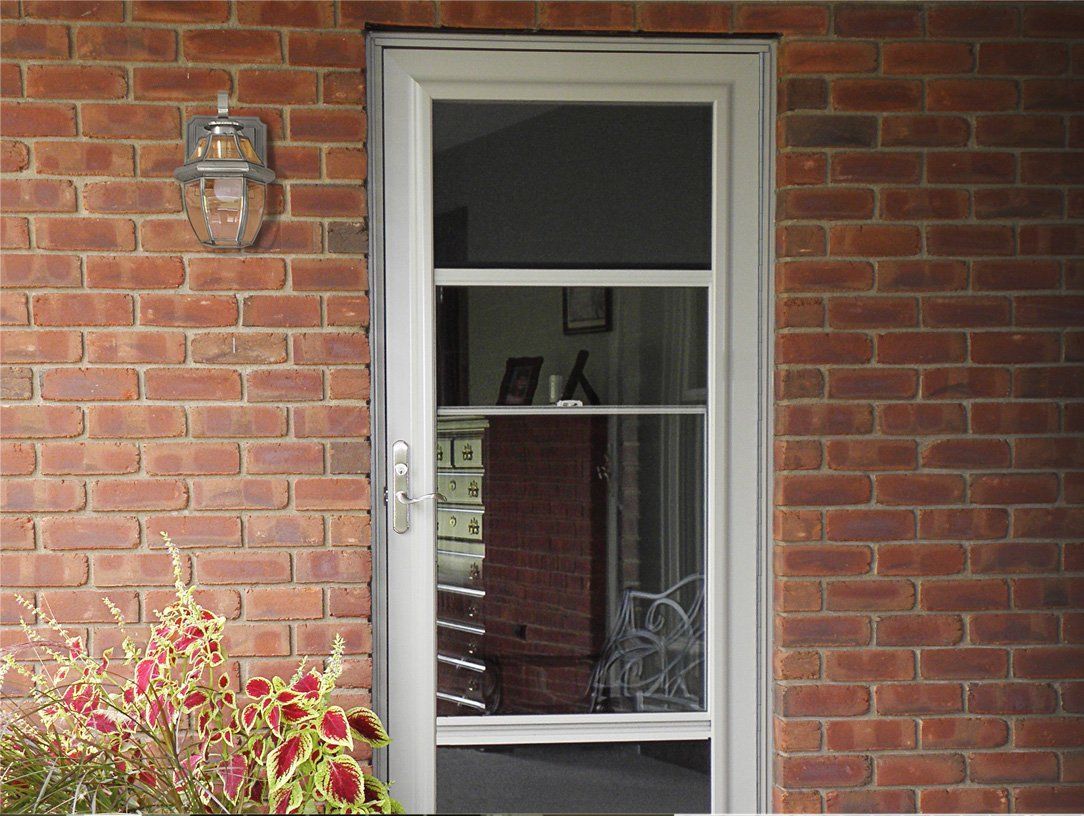 White framed screen door with three glass panels set in a red brick wall, with a decorative light fixture and plants nearby.