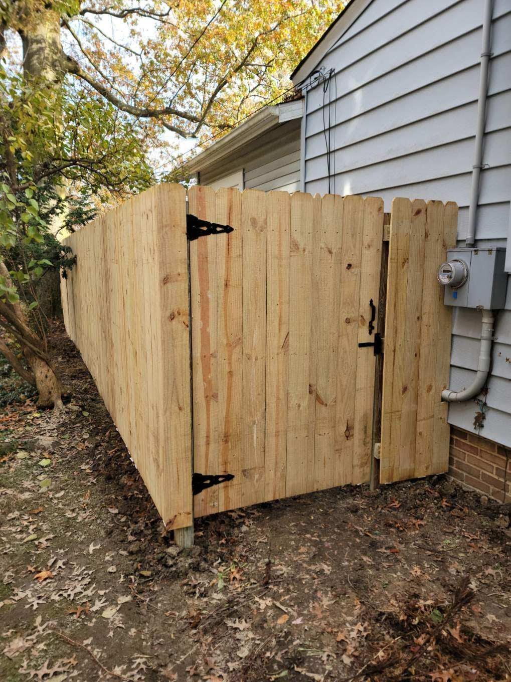 Wooden fence with gate next to a house; partially obscuring an electrical box; brown, outdoors.
