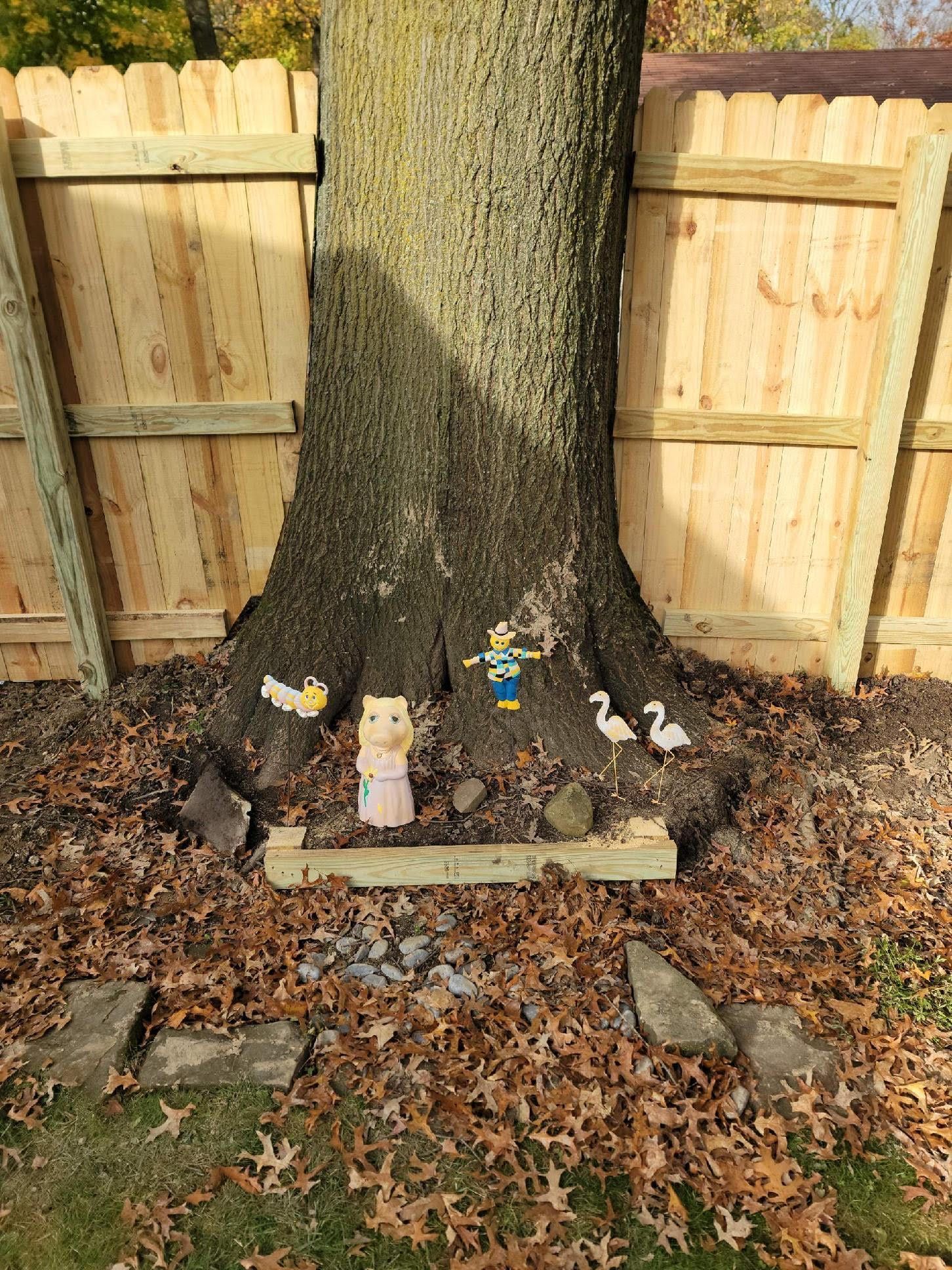 Tree trunk with small figurines, surrounded by fallen leaves, a fence in the background.