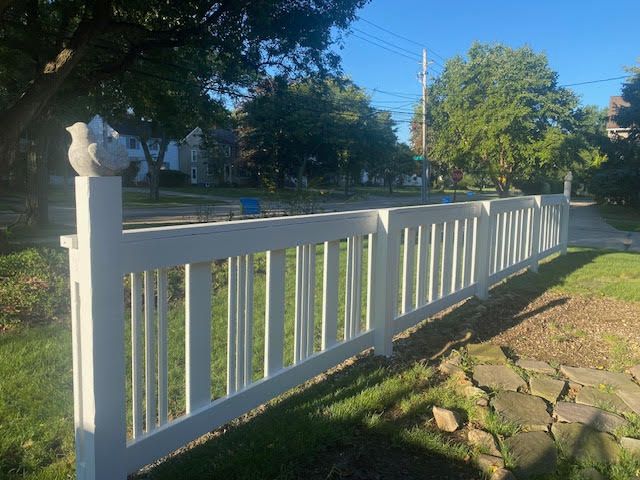 White picket fence with decorative bird toppers lining a grassy yard, with a street and trees in the background.