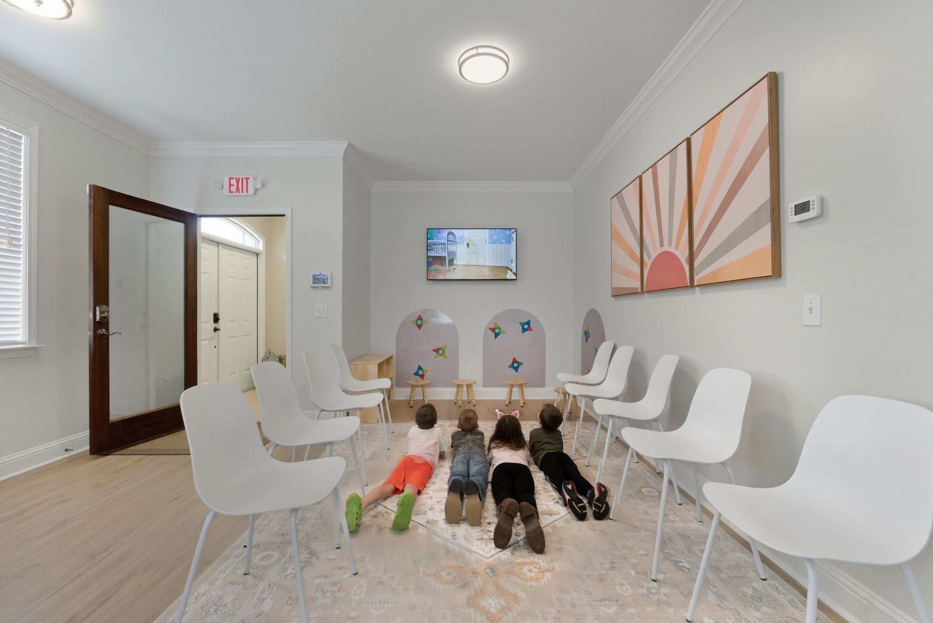 Four children lie on a rug, watching a wall-mounted television in a brightly lit waiting room with white chairs.