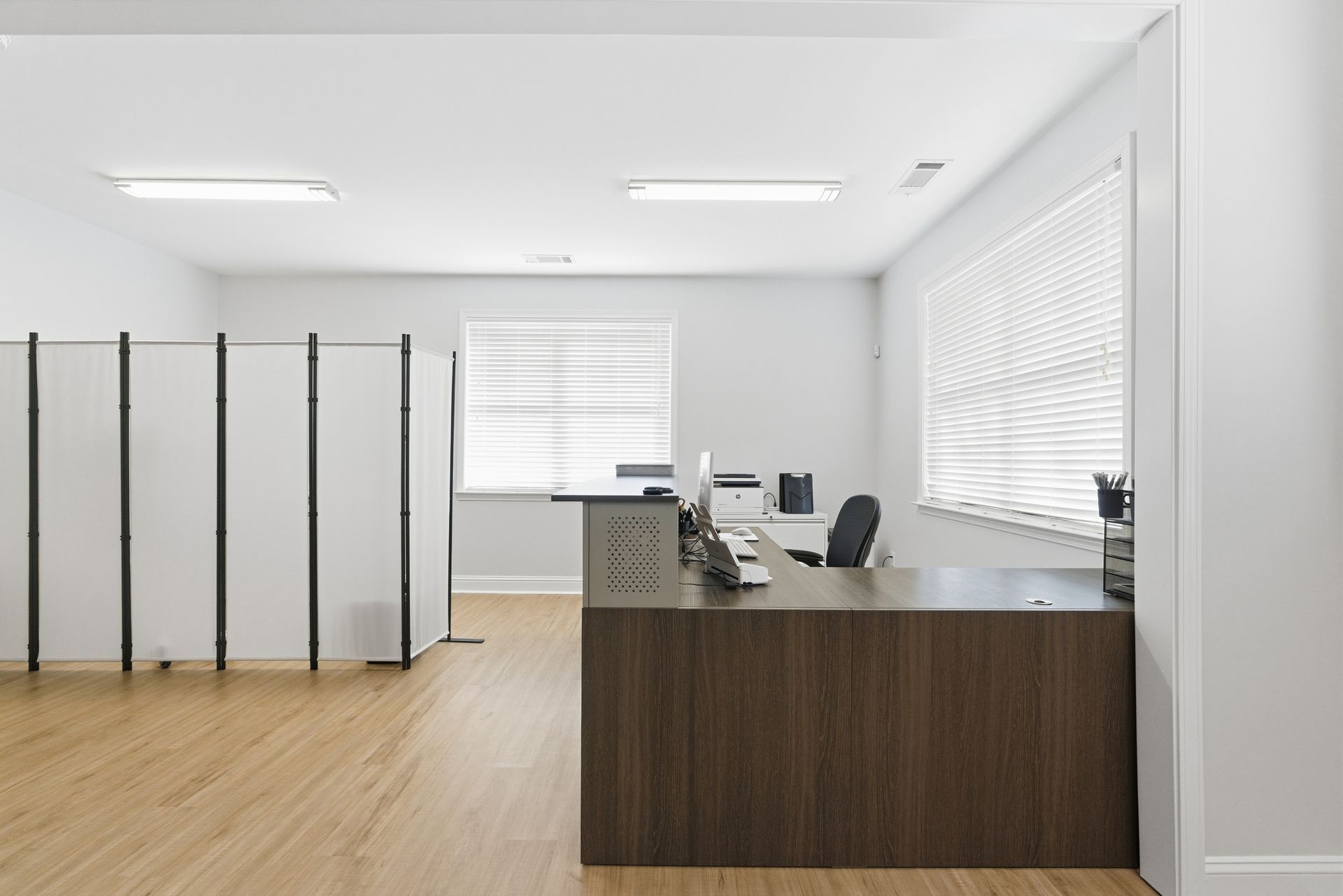 Modern reception office with a dark wood desk, light wood floors, white walls, room dividers, and window blinds.