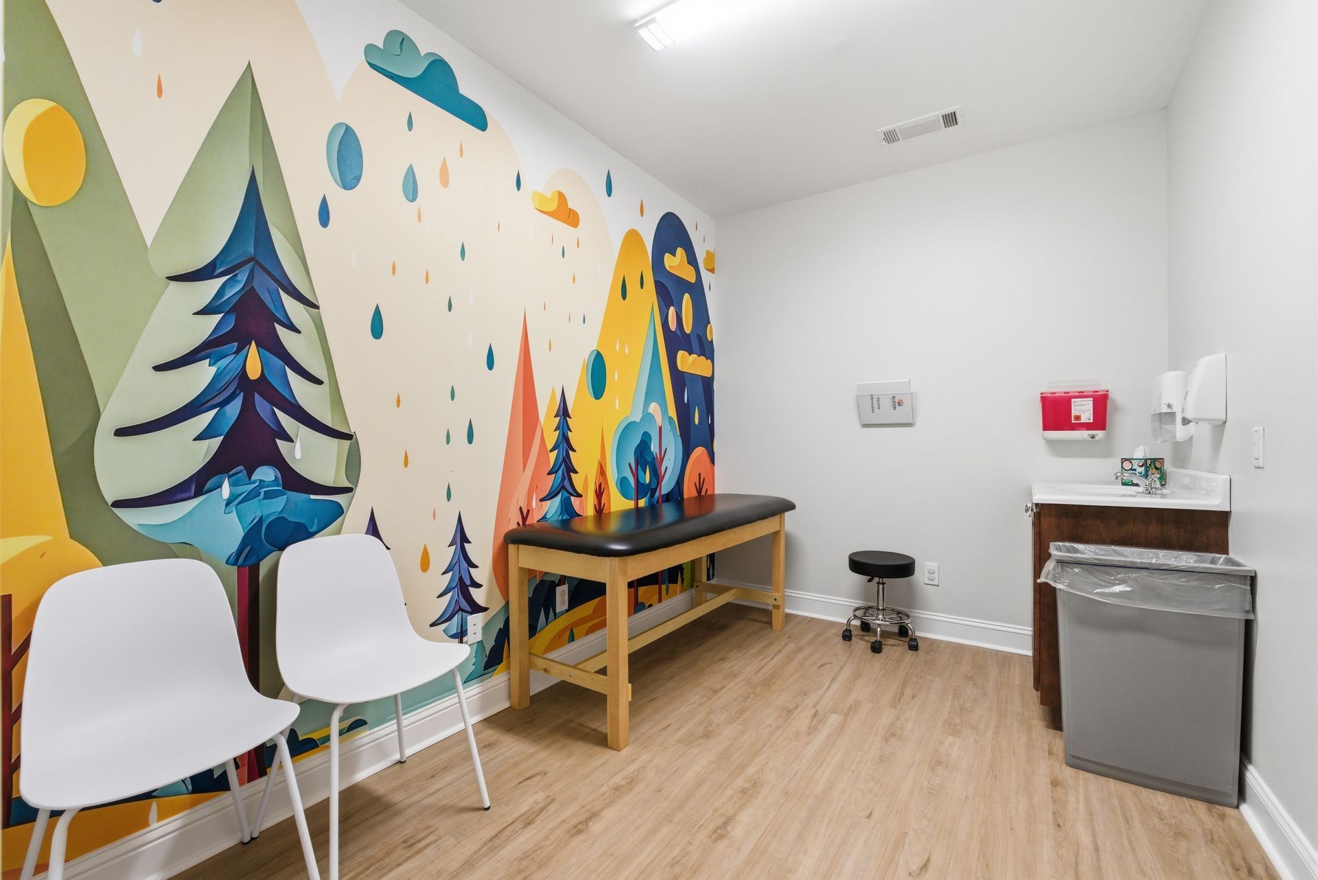 A clinic exam room with a colorful mountain mural, exam table, two white chairs, a rolling stool, and a trash bin.