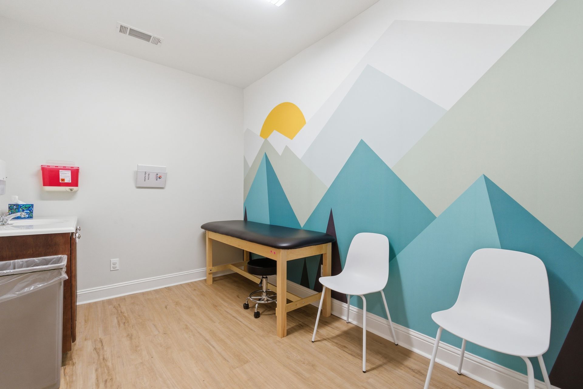 A medical examination room with a wooden exam table, two white chairs, and a mountain-themed wall mural.