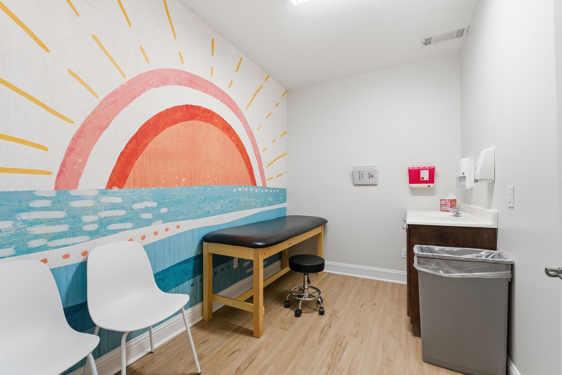 A medical exam room with white chairs, an exam table, a stool, and a colorful sun mural on the wall.