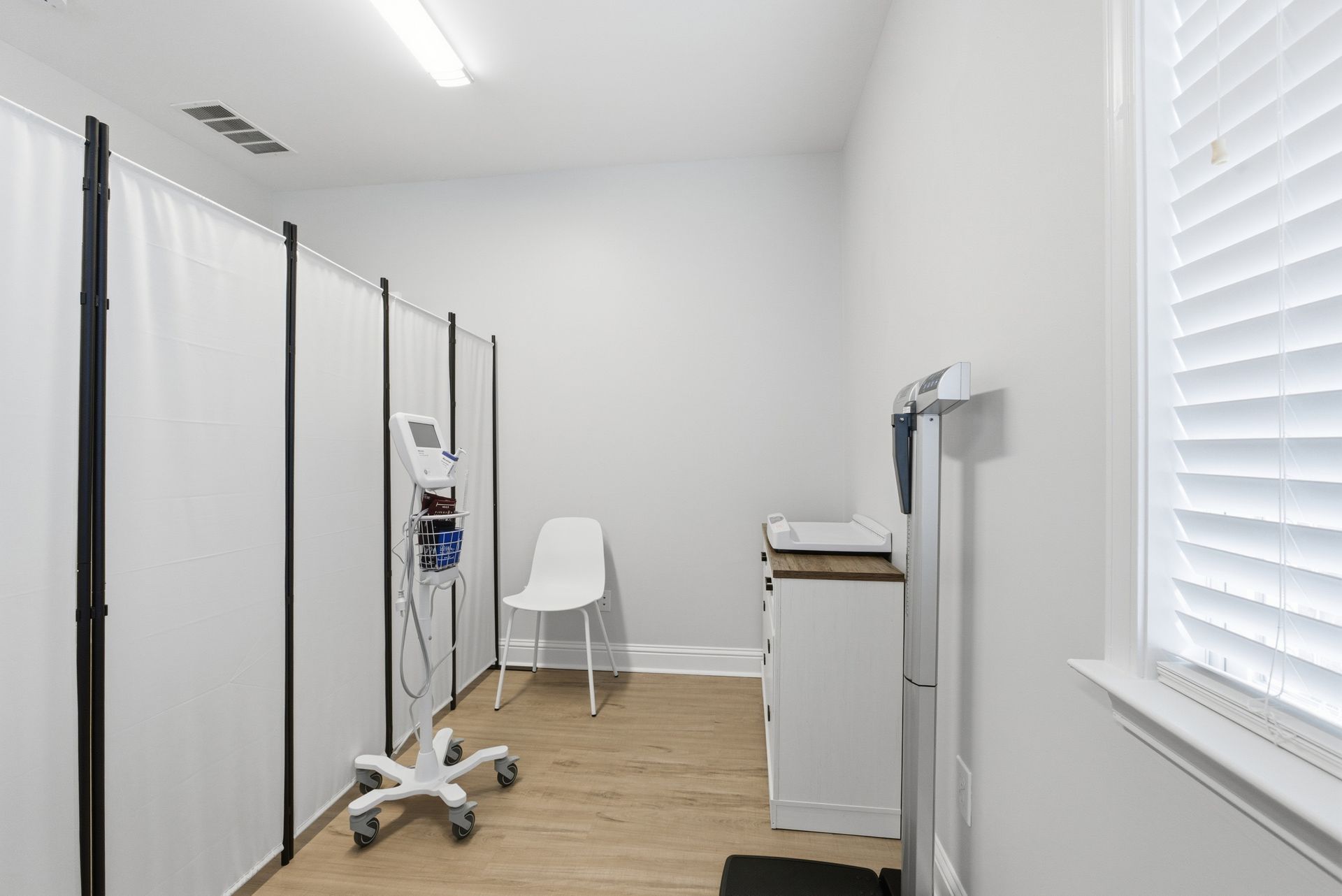 A sterile medical examination room with light gray walls, a white privacy screen, a mobile medical device, and a chair.
