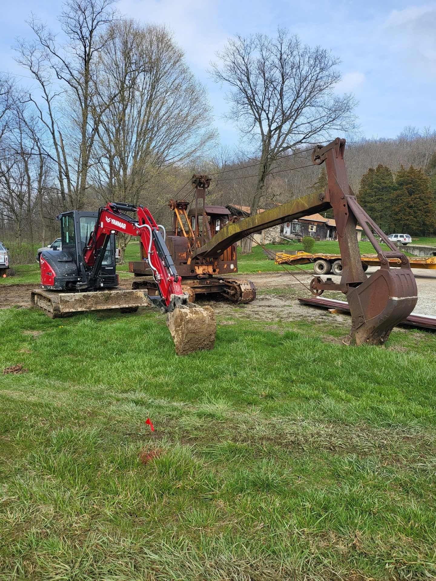 Mini excavator and tractor moving dirt in a grassy field near trees under a cloudy sky