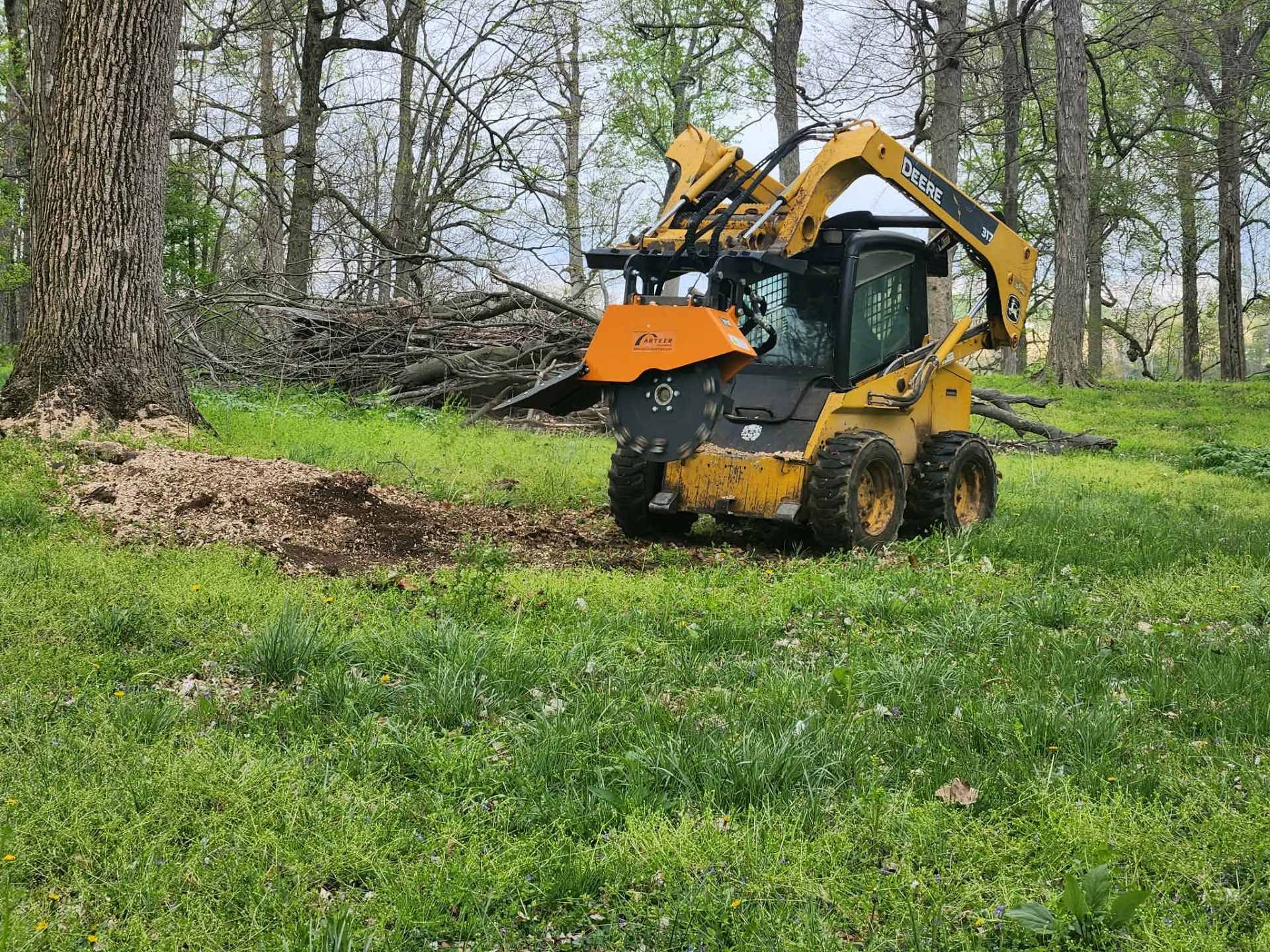 Yellow excavator with raised arm in a grassy wooded area near a pile of branches