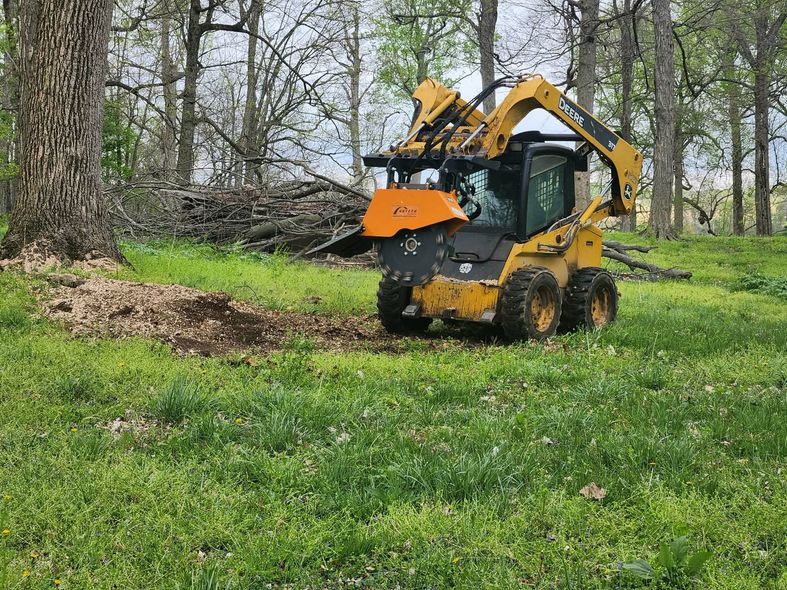 Yellow excavator in a grassy wooded area, lifting a pile of dirt and branches.