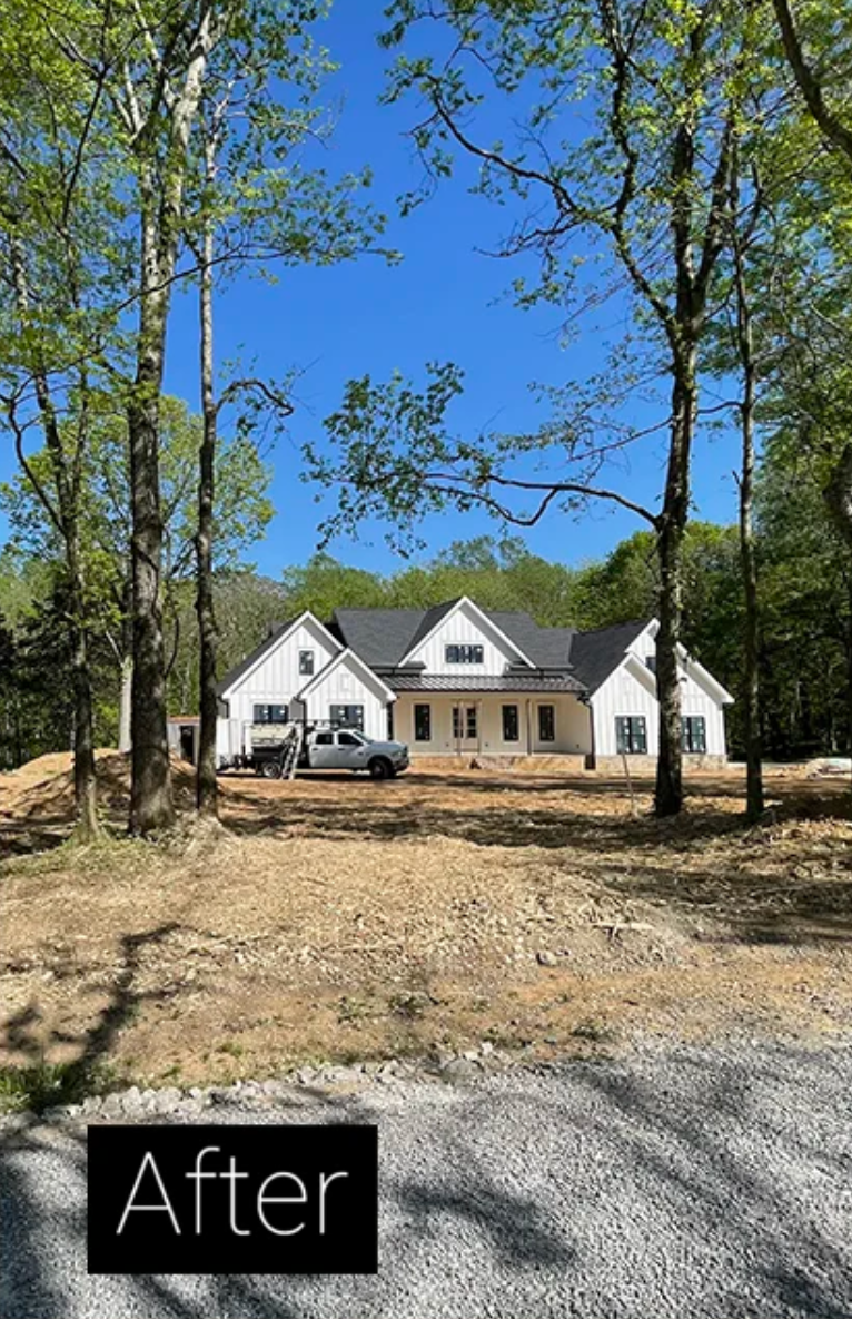 New house with white siding and tan brick behind bare yard, framed by trees under a blue sky.