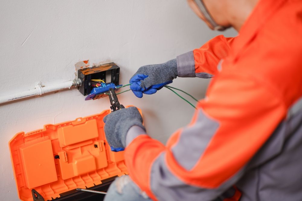 Electrician in orange coveralls and gloves wiring electrical box on a wall.