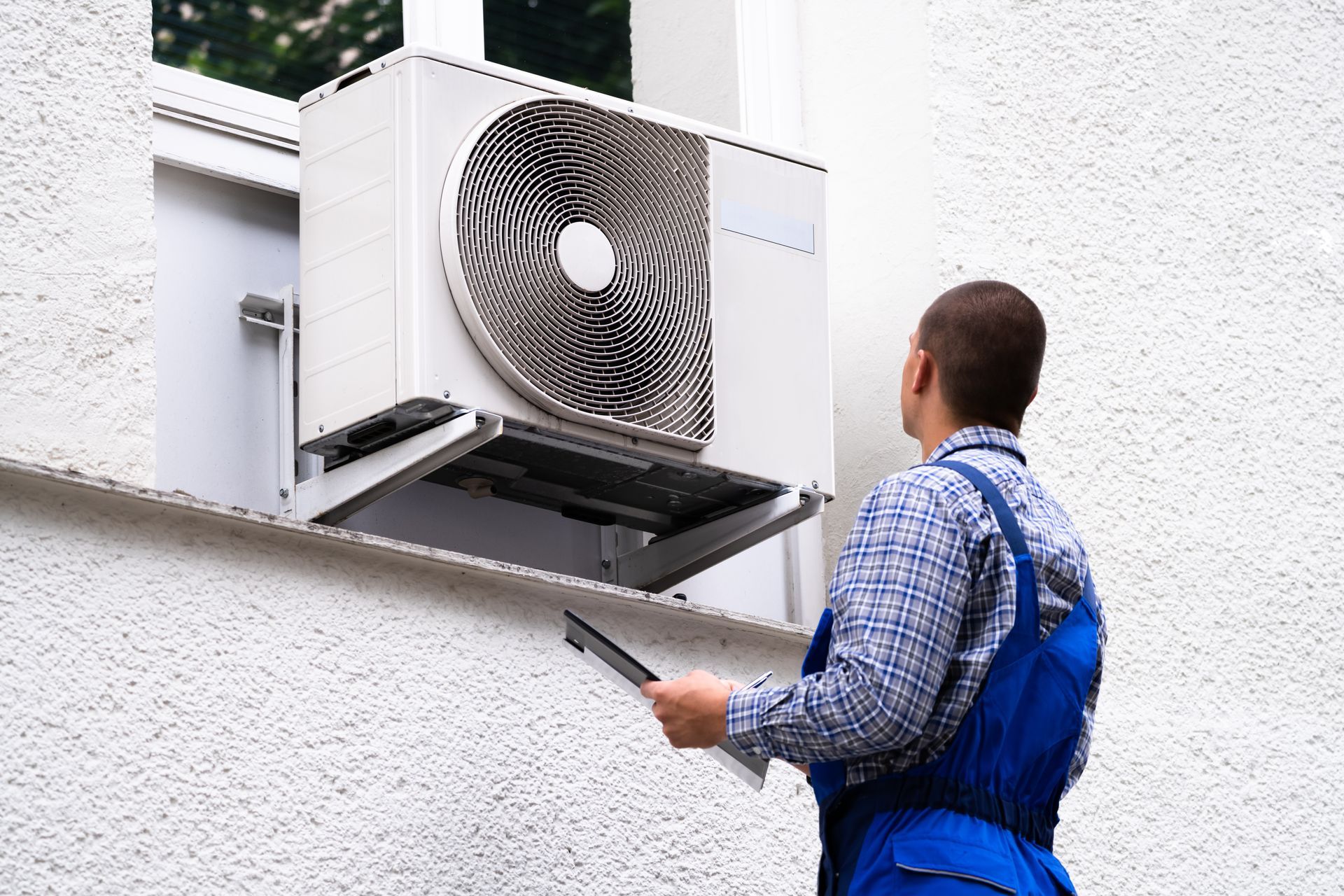 A man in overalls examines an air conditioner while holding a clipboard.