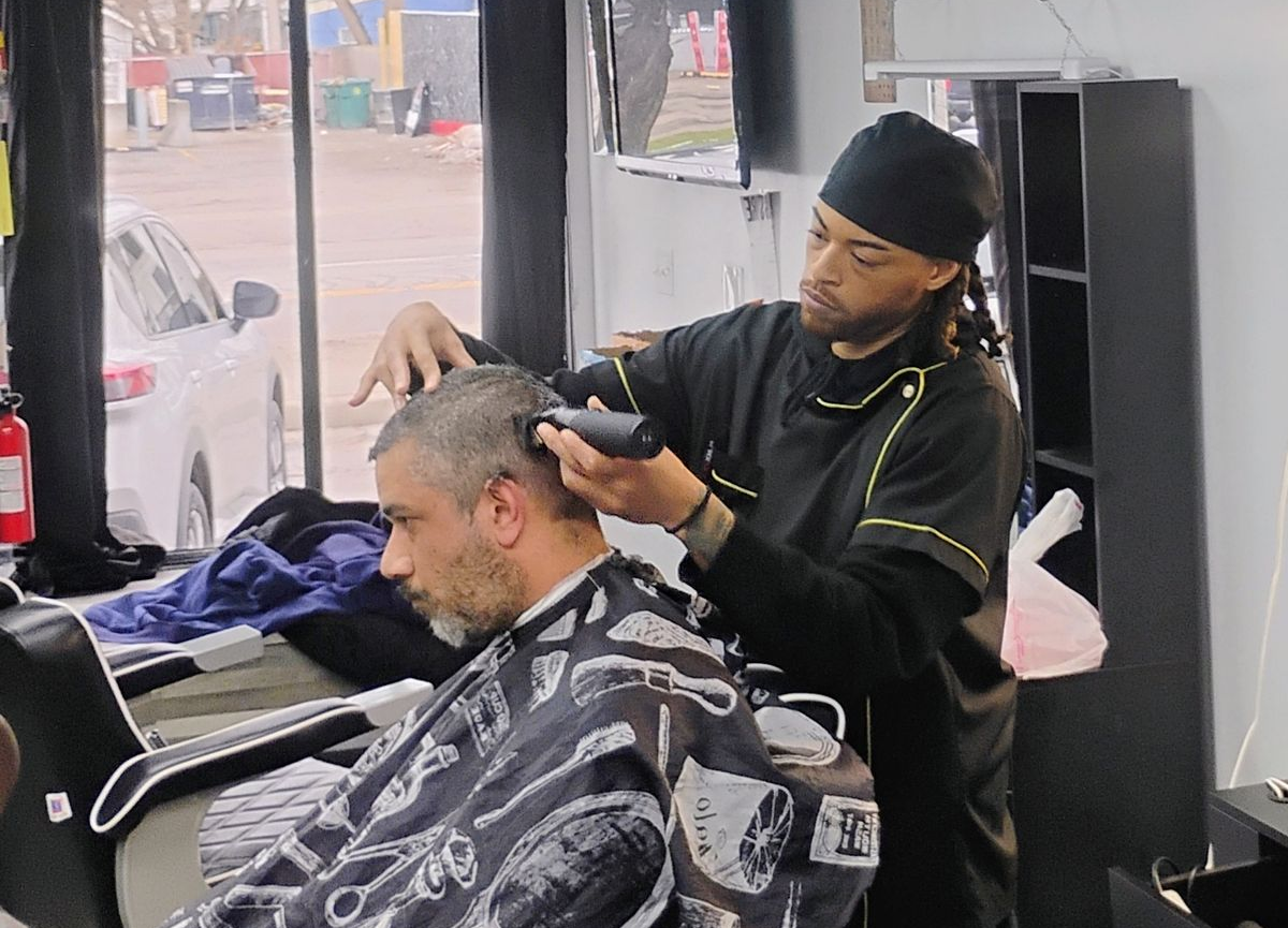 A barber in a black uniform uses electric clippers to cut the hair of a customer sitting in a chair wearing a barber cape.