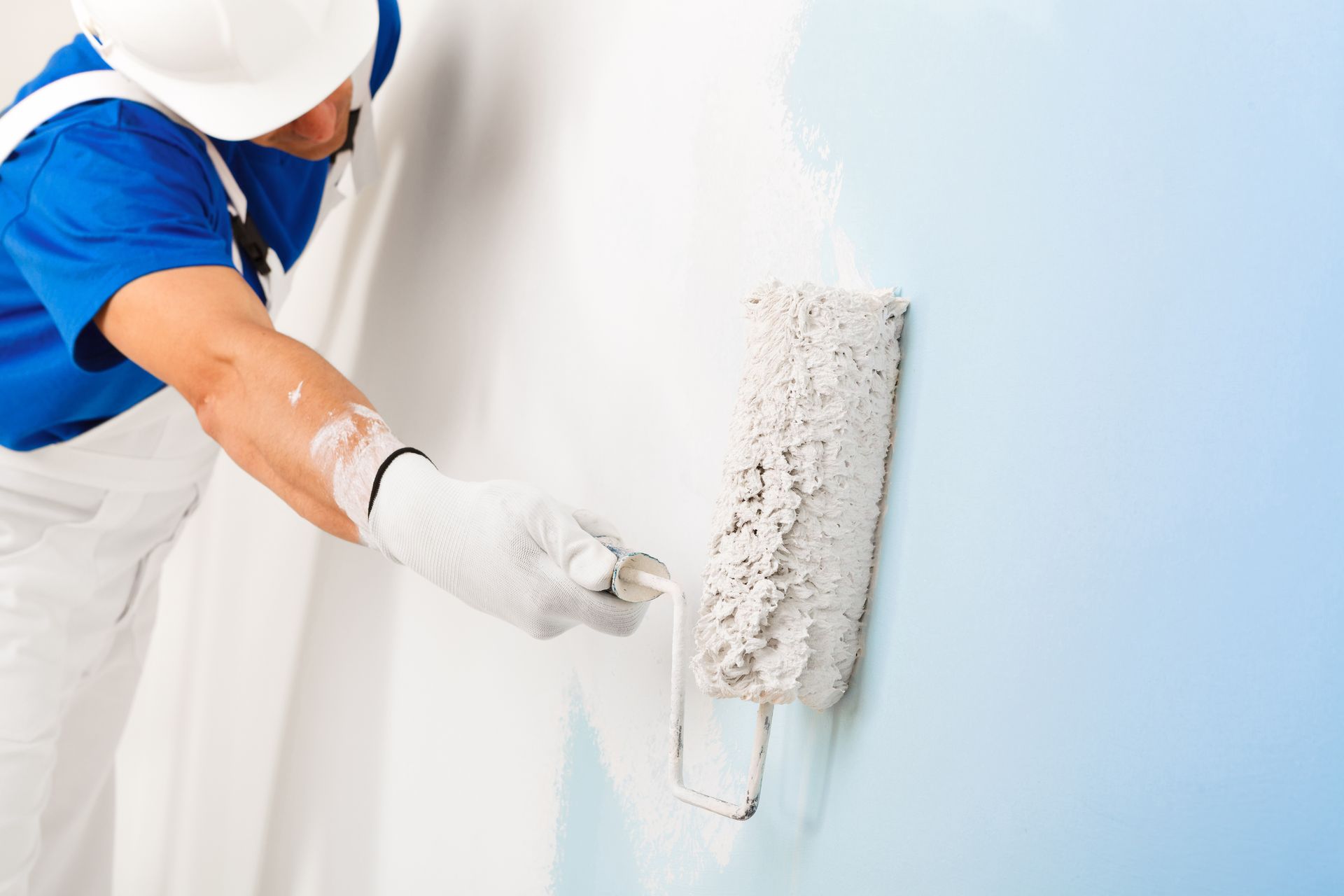 Person painting a white wall with a roller, wearing blue shirt and white gloves