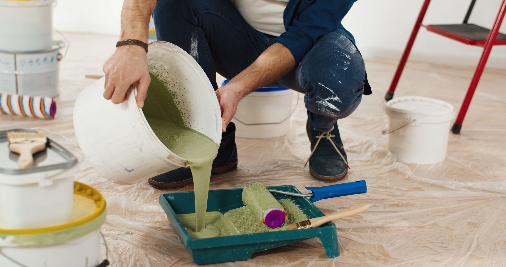 Person pours green paint into a tray while painting a room floor, with tools and buckets nearby.