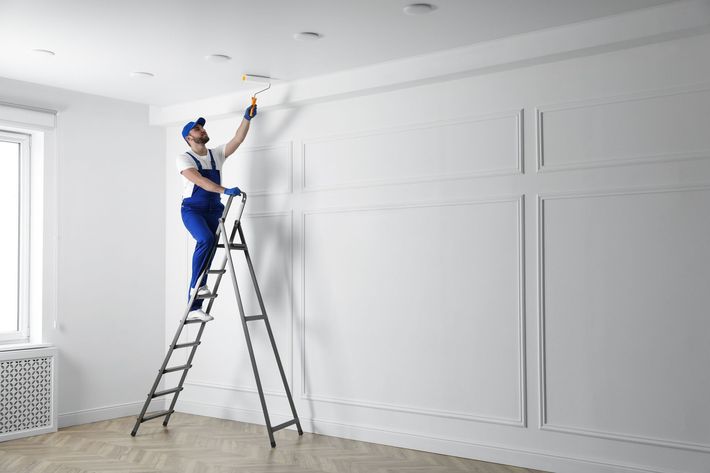 Worker on a ladder painting a white ceiling in a bright empty room.
