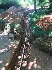 A long trench dug in a yard with a white pipe running through it, surrounded by dirt and stone walls.