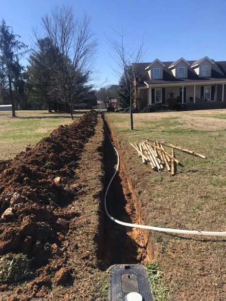 Trench dug in a yard, with buried conduit, leading towards a house with a porch and two trees.