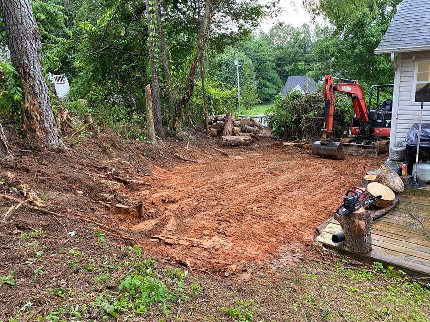Red dirt area cleared by excavator next to a house and trees. Logs and debris are visible.