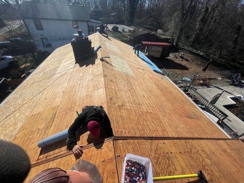Roofers working on a house roof on a sunny day. Person in red cap kneels, installing roofing materials.