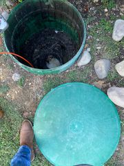 Open green septic tank, lid off, revealing dark contents. A person's boot and jeans are visible.