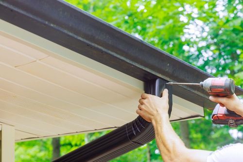 Person installing a black gutter with a power drill on a house with beige siding.