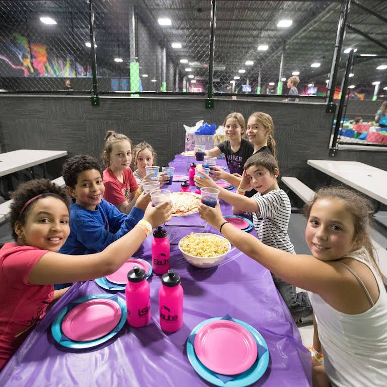 Kids at a party table cheering, with pizza and drinks. Party is set in a trampoline park.