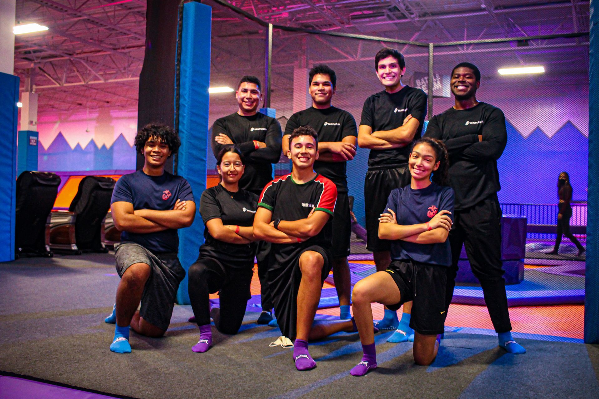 A group of eight people, arms crossed, pose in a trampoline park. They wear black shirts with a logo and blue/purple socks.