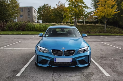 Blue BMW M2 parked in an asphalt parking lot with a building and trees in the background.