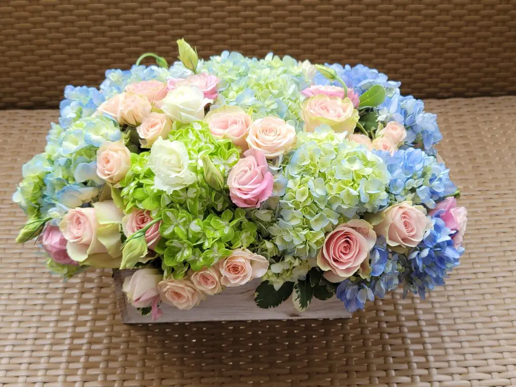 A white box filled with pink and blue flowers is sitting on a wicker table.