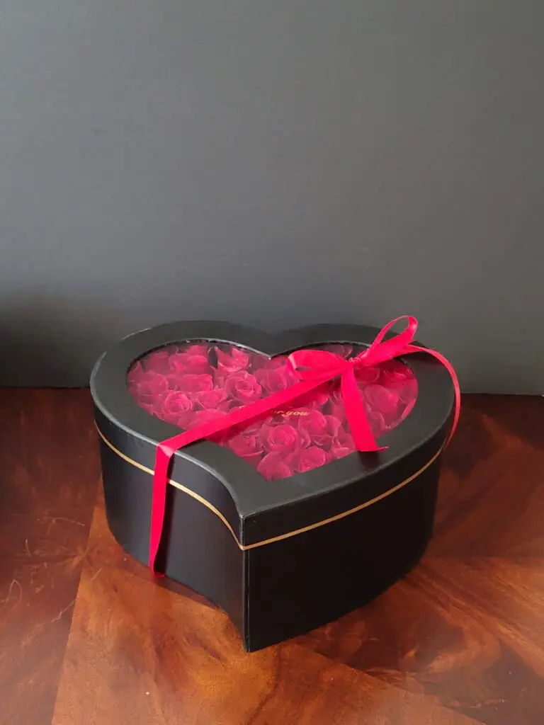 A heart shaped box filled with red roses is sitting on a wooden table.
