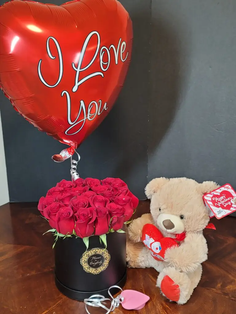 A teddy bear is sitting next to a box of roses and a heart shaped balloon that says i love you.