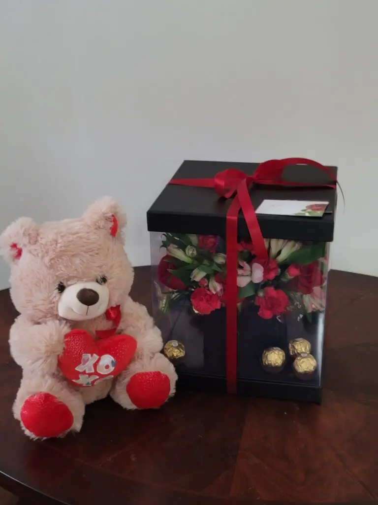 A teddy bear holding a heart next to a box of flowers