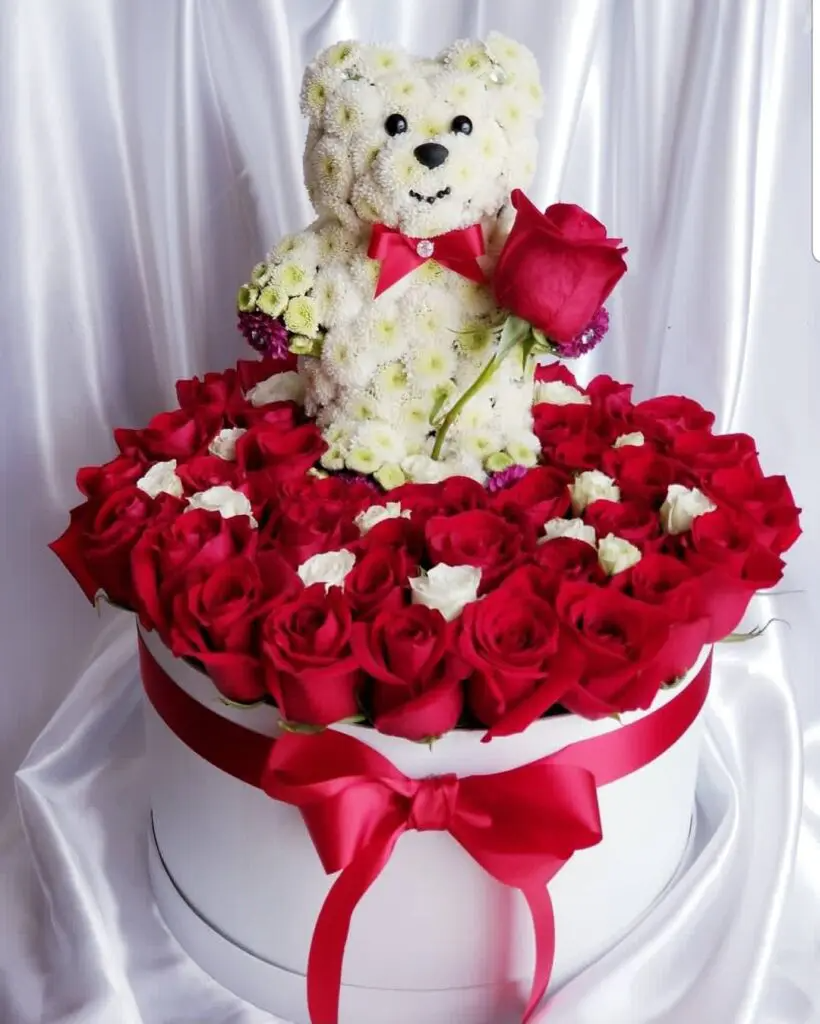 A white teddy bear is sitting on top of a box of red roses