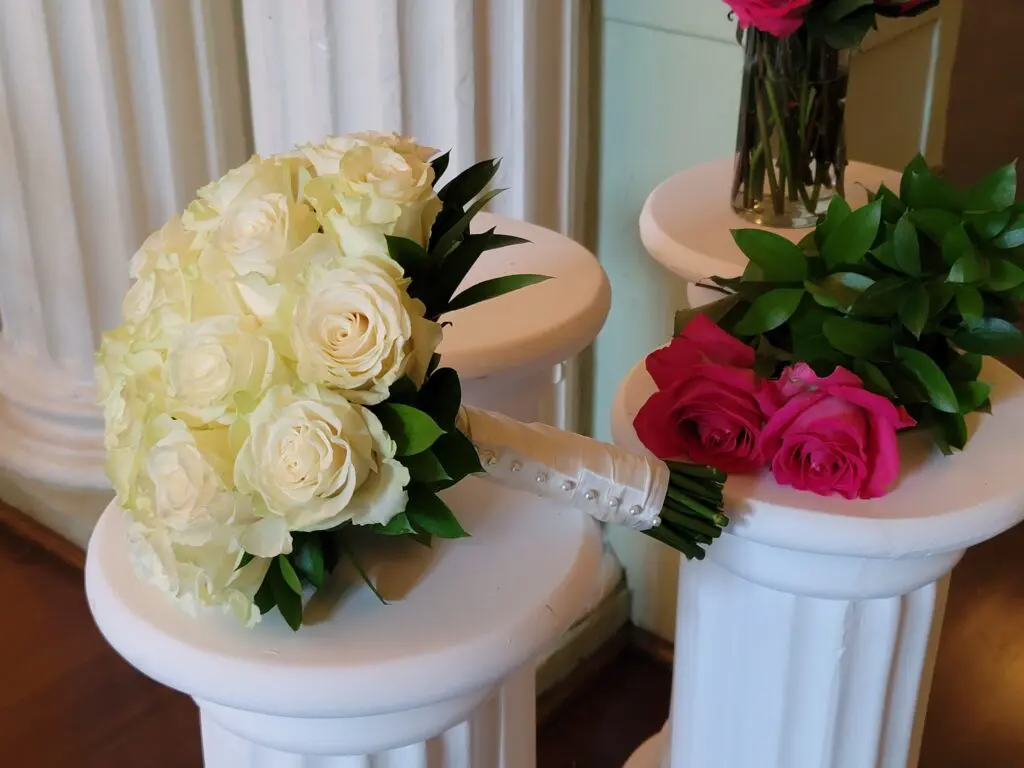 A bouquet of white roses sits on a white pedestal next to a vase of pink roses