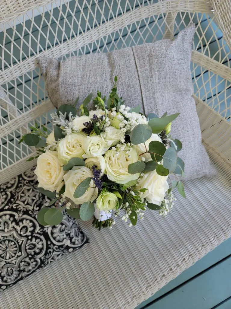 A bouquet of white roses is sitting on a wicker bench next to a pillow.