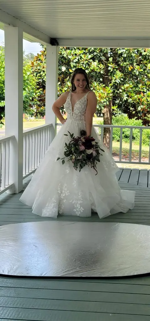 A woman in a wedding dress is standing on a porch holding a bouquet of flowers.