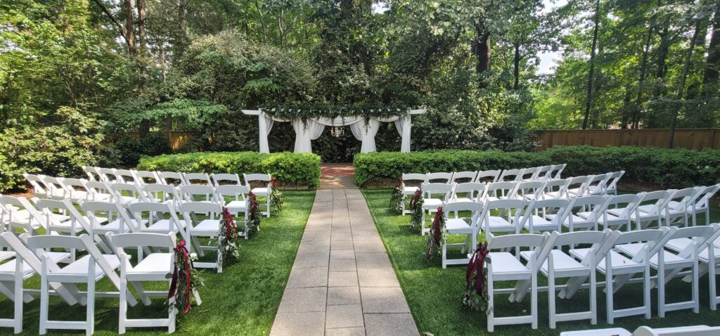 A row of white chairs are lined up in a garden for a wedding ceremony.