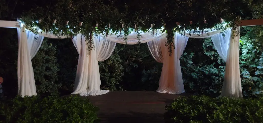 A pergola with white curtains and lights is lit up at night.