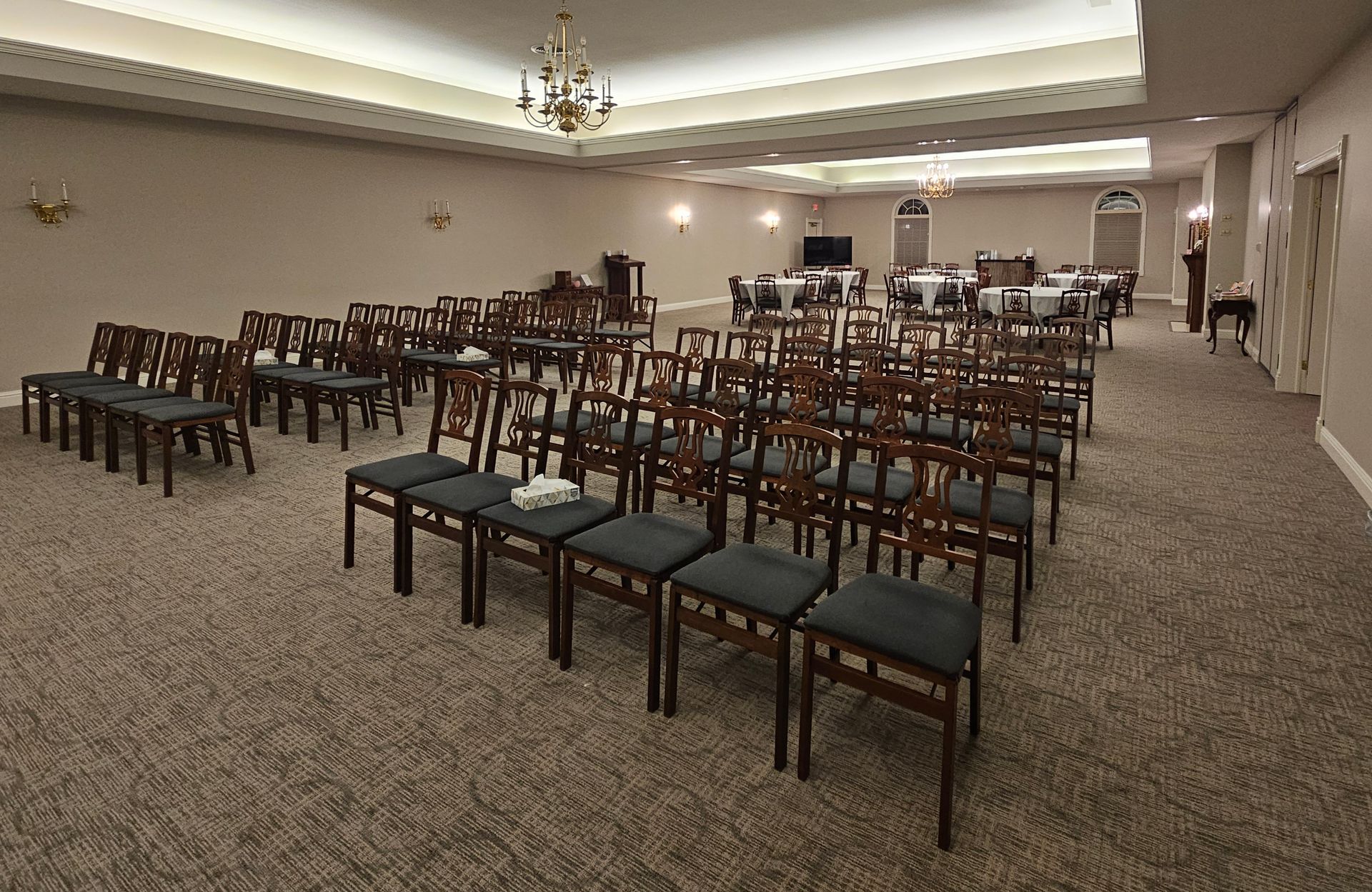 Empty banquet hall with rows of dark chairs and tables on beige carpet under chandeliers