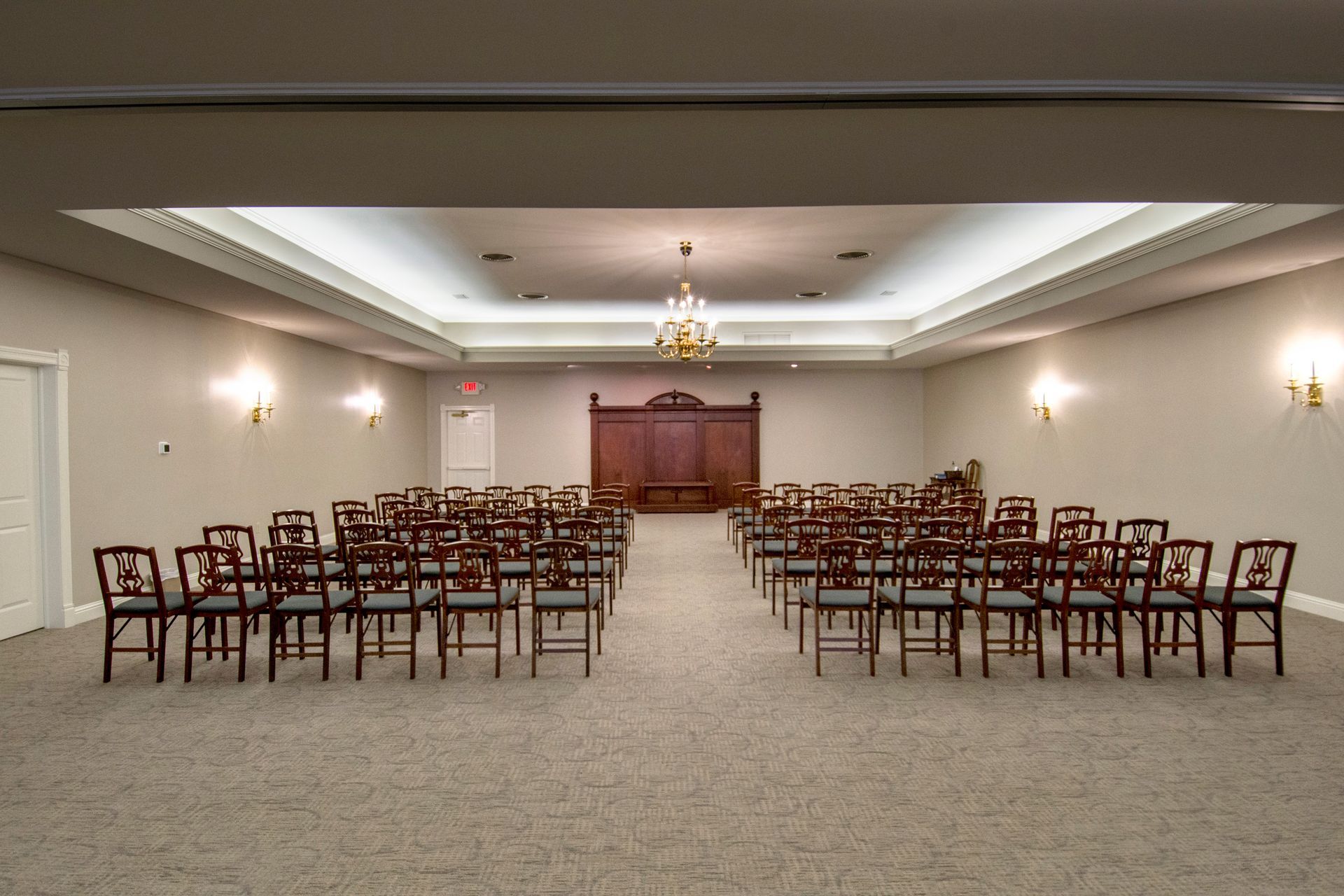 Rows of empty chairs face a wooden structure in a carpeted room with a chandelier.
