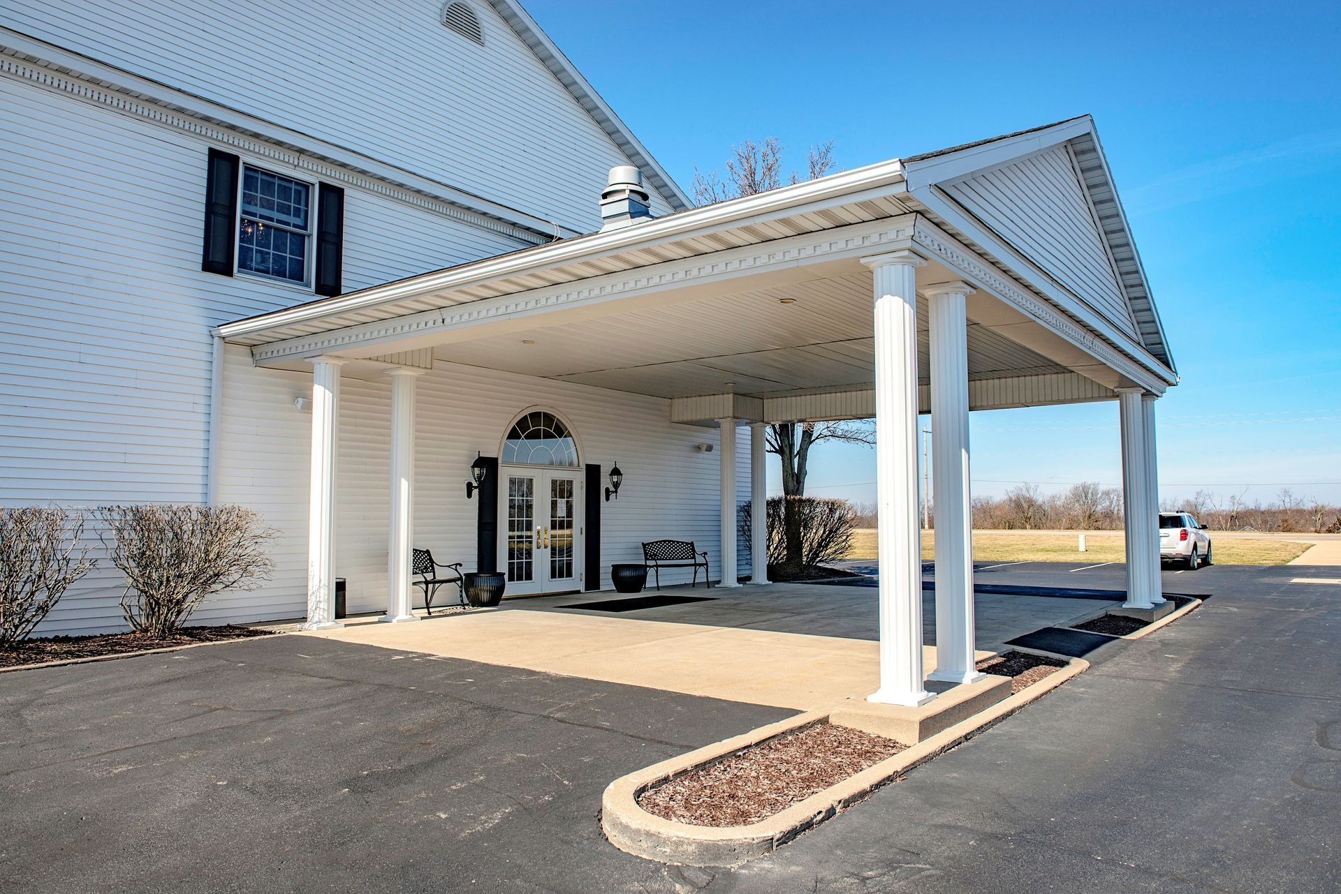 White building with a covered entrance, columns, and a paved driveway. Blue sky in the background.