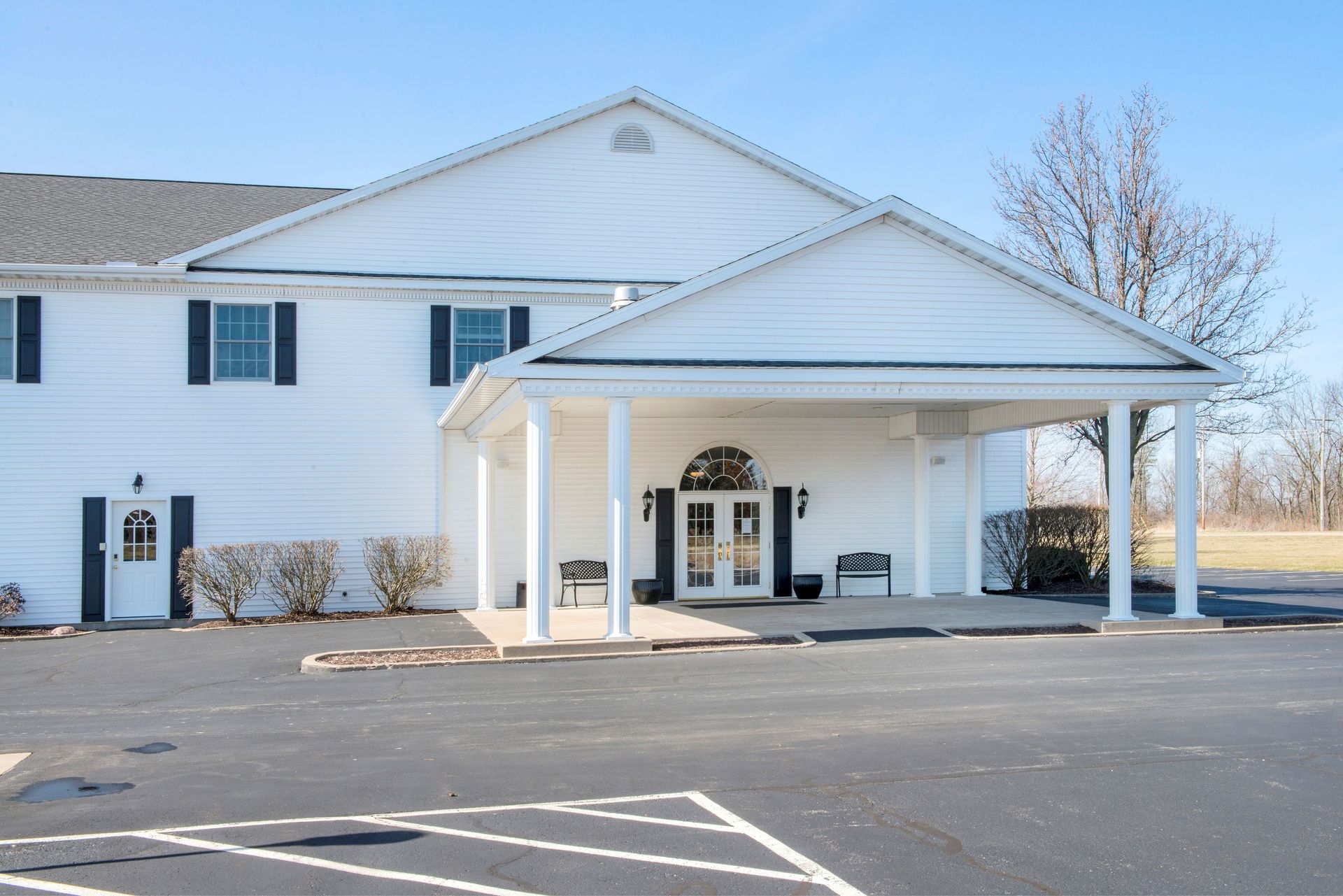White building with columns and black roof, entrance under a portico, in a parking lot on a sunny day.