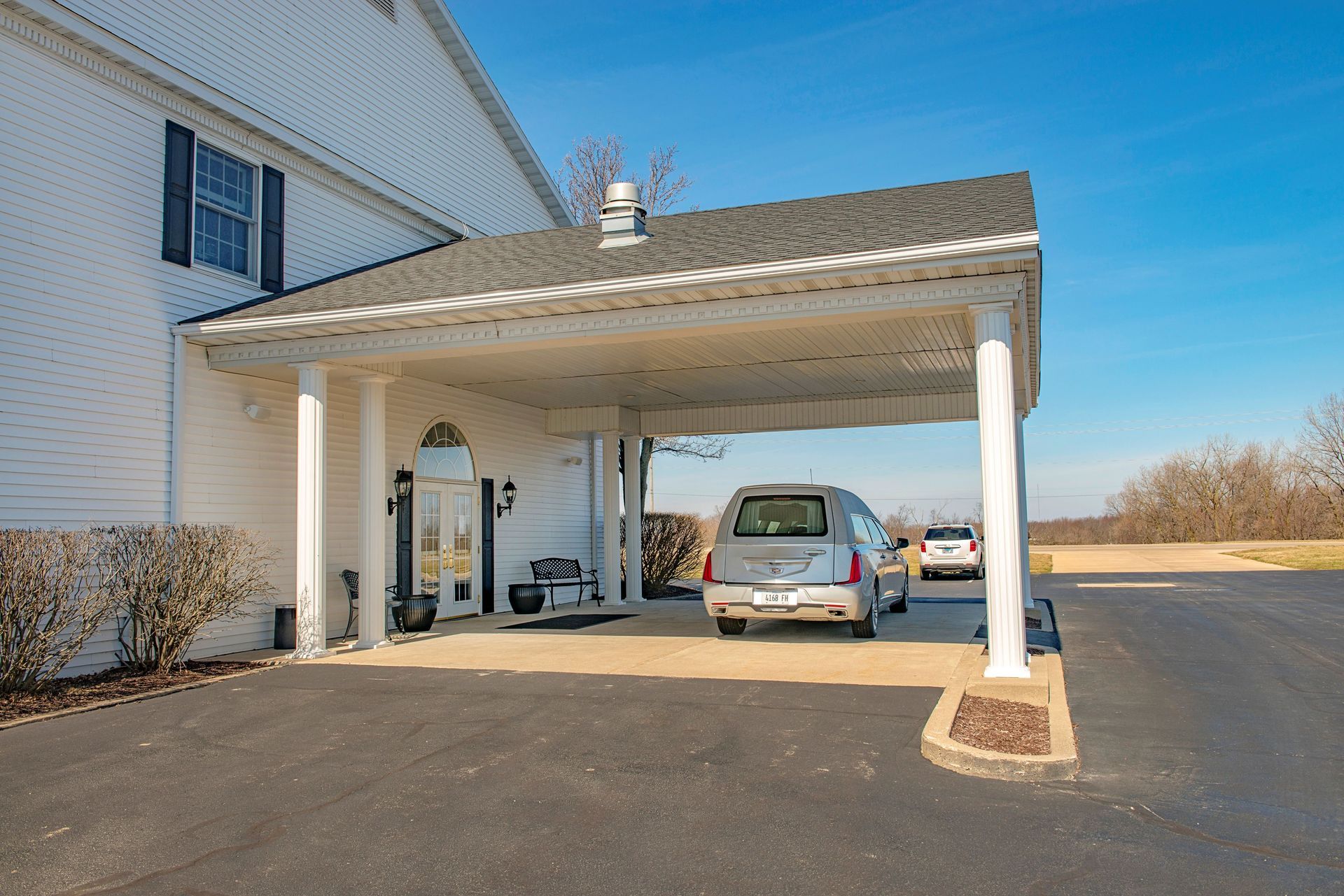 A hearse parked under a white covered entrance to a white building with a blue sky.