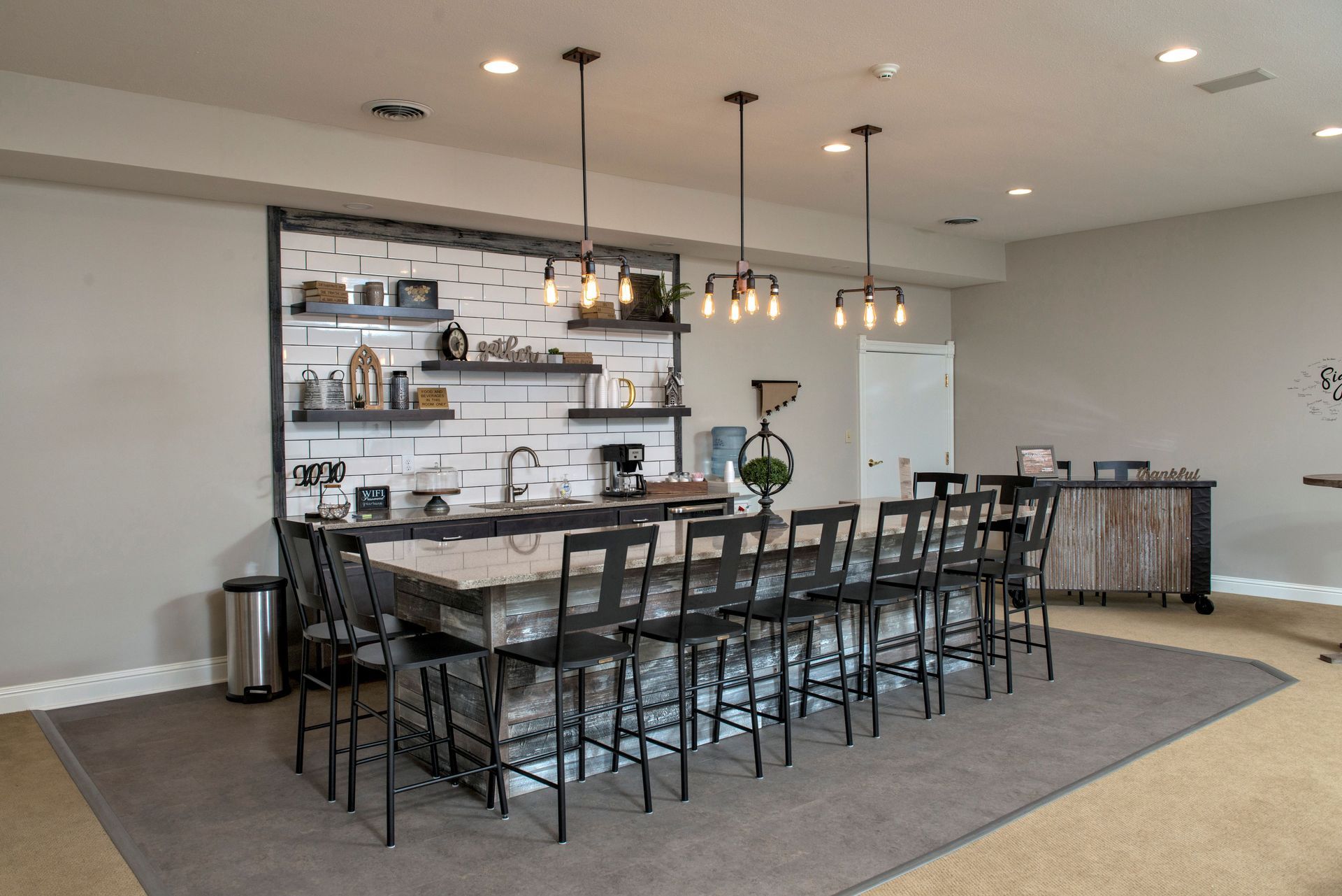Modern kitchen/bar with long island, stools, shelves, pendant lights. White brick accent wall and gray rug.