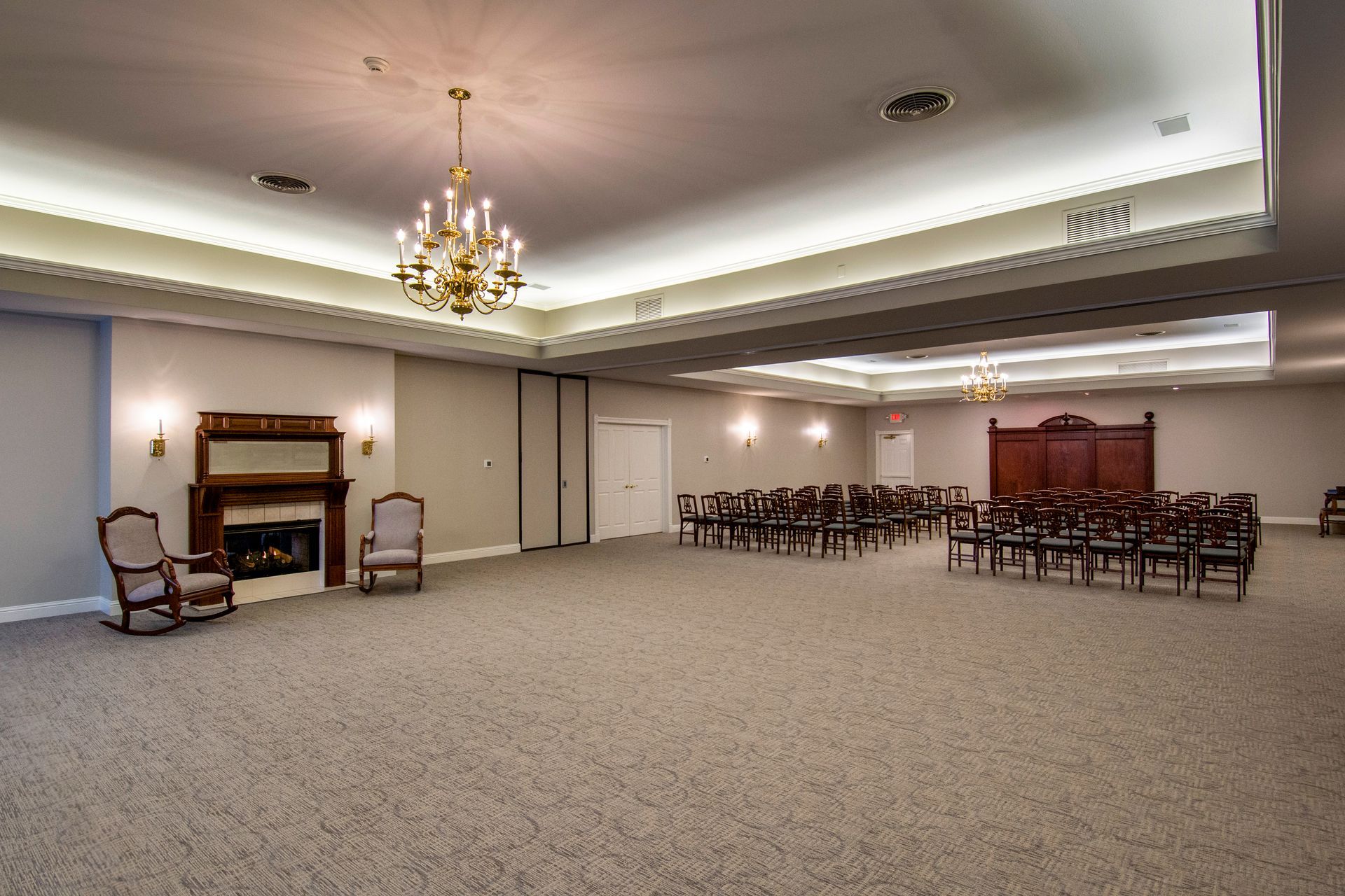 Large, empty room with chairs arranged in rows; fireplace and ornate chandelier present.